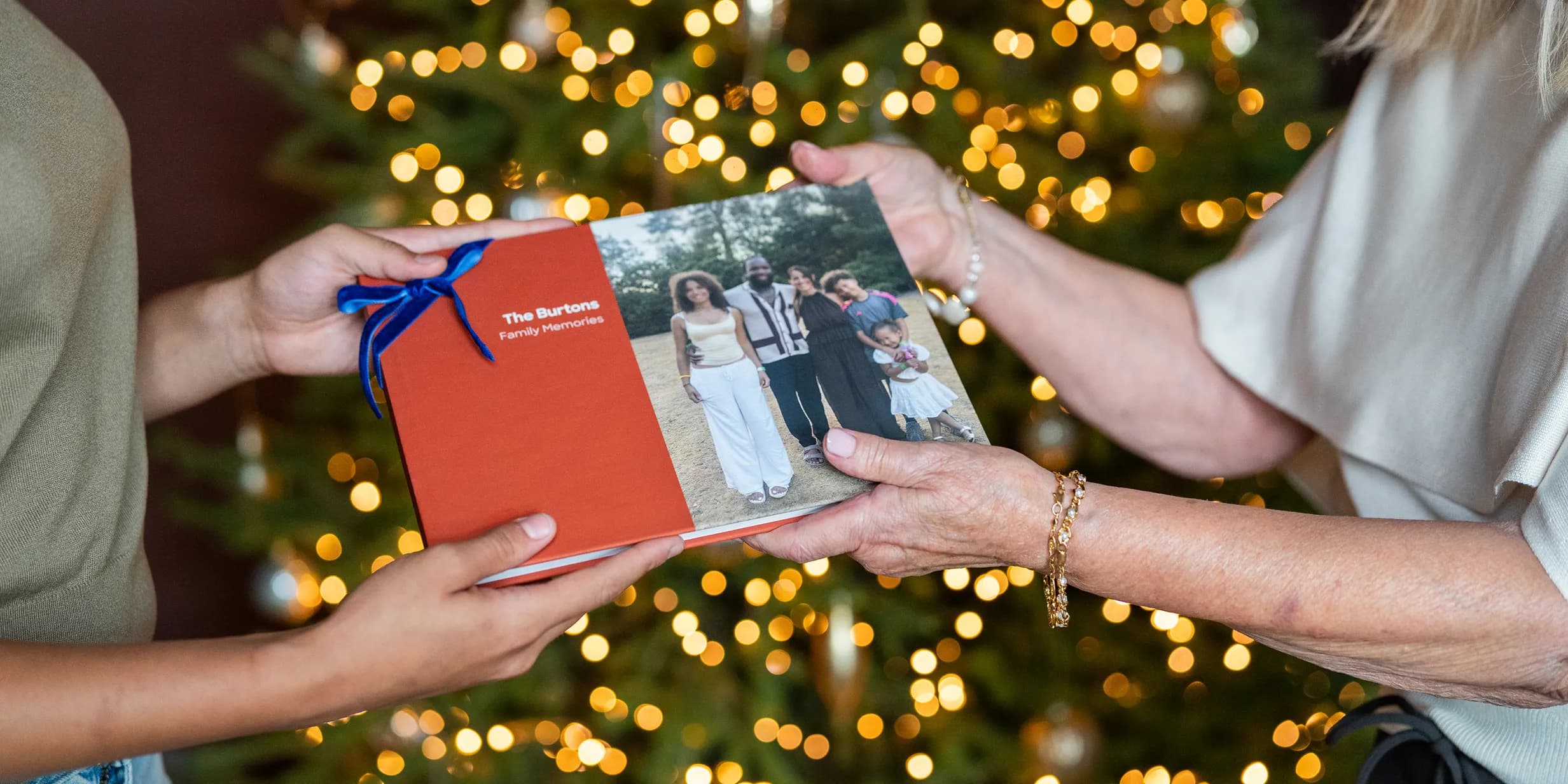Two people exchanging a family photo album with a Christmas tree in the background.