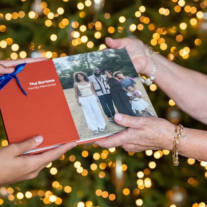 Two people exchanging a family photo album with a Christmas tree in the background.