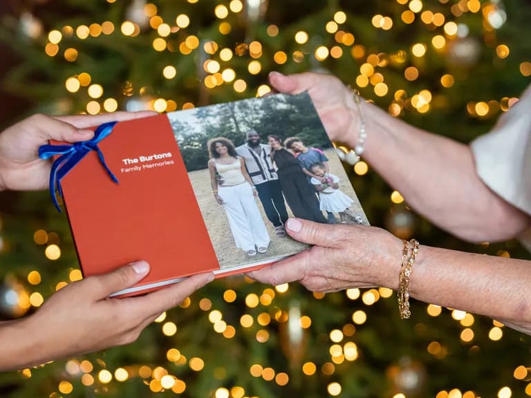 Two people exchanging a family photo album with a Christmas tree in the background.