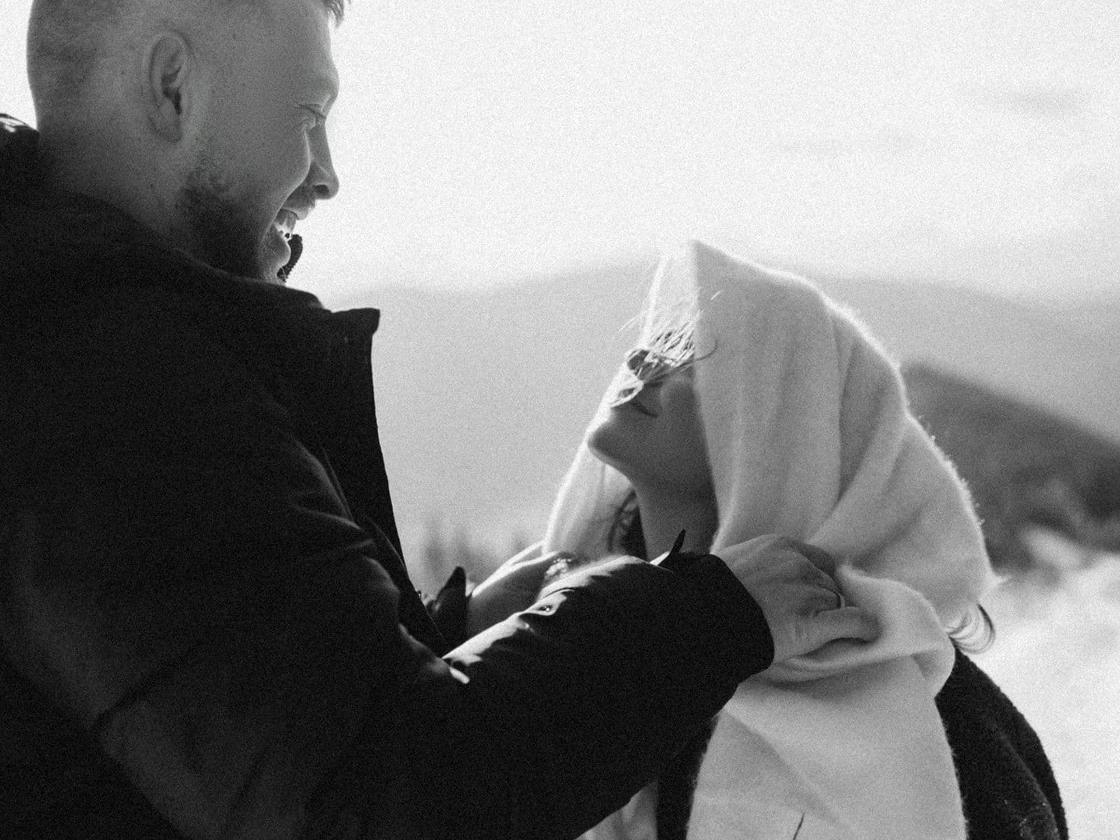 A man and woman smiling at each other on a snowy mountain trail. The woman is wrapped in a blanket. Black and white photo.