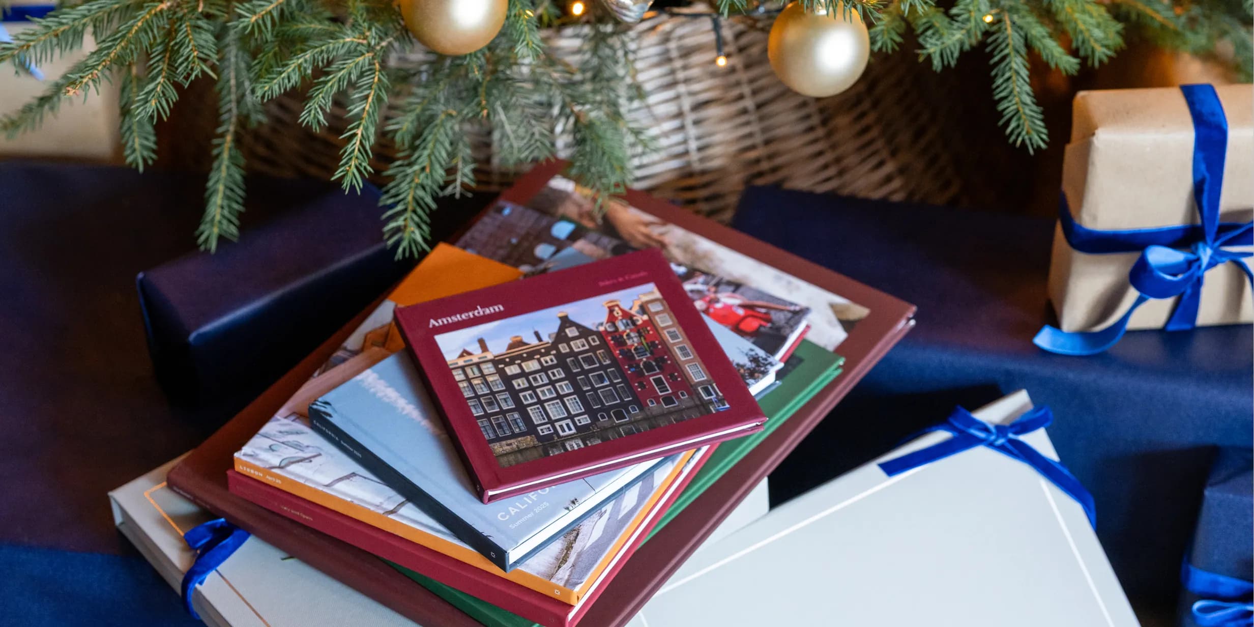 Stack of travel photo albums, including "Amsterdam", under a Christmas tree with gold ornaments, surrounded by gifts with blue ribbons.