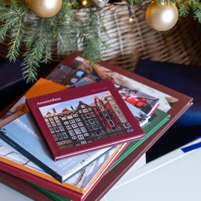 Stack of travel photo albums, including "Amsterdam", under a Christmas tree with gold ornaments, surrounded by gifts with blue ribbons.