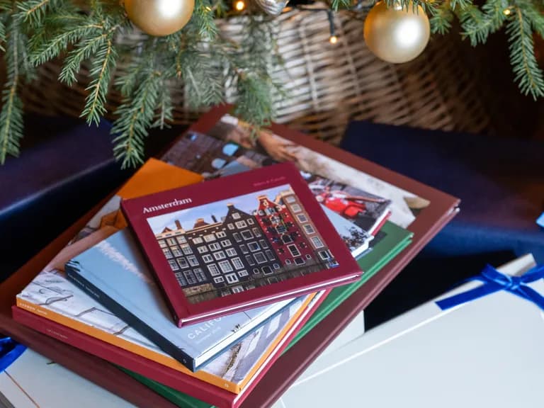 Stack of travel photo albums, including "Amsterdam", under a Christmas tree with gold ornaments, surrounded by gifts with blue ribbons.