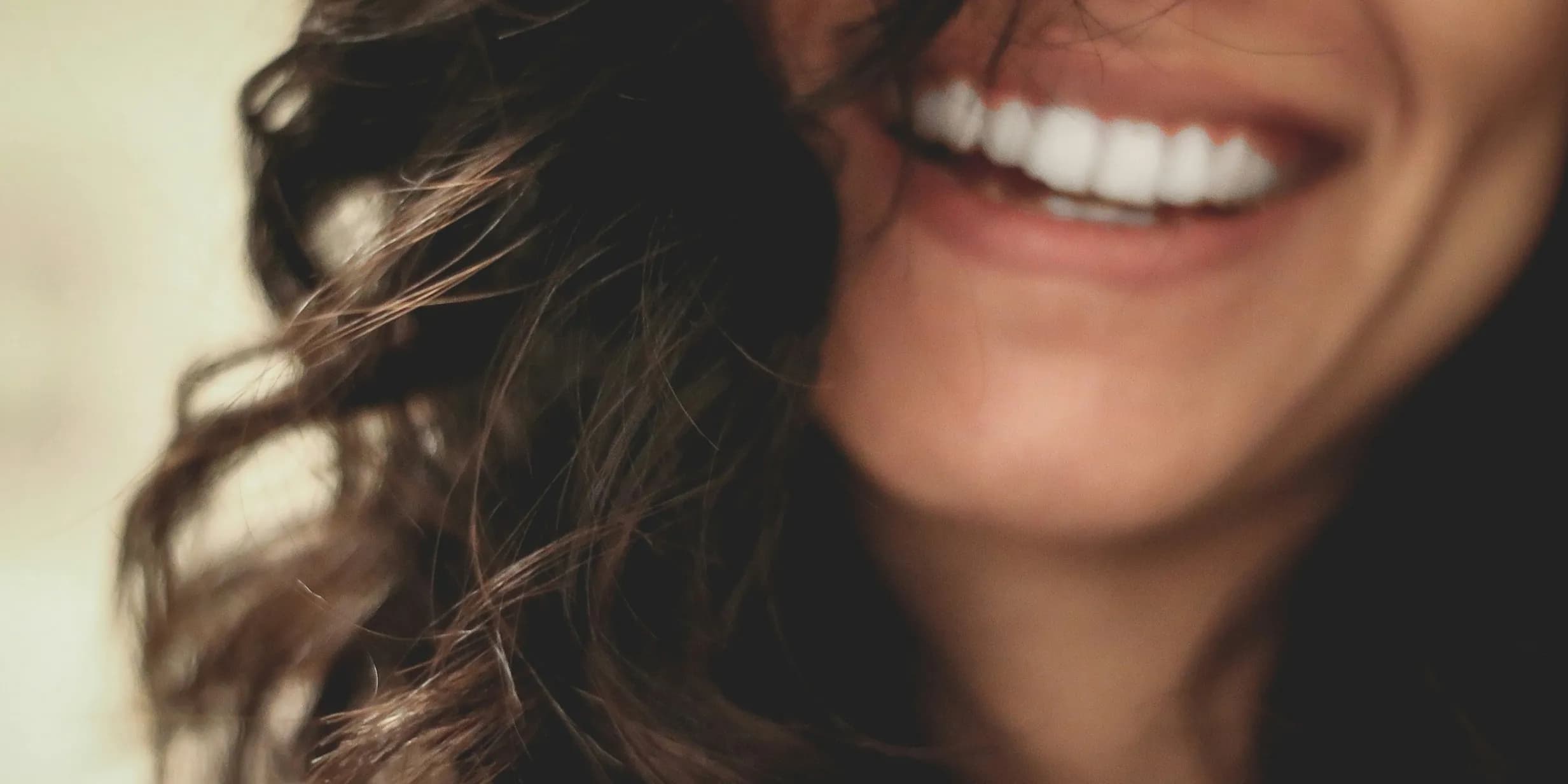Close-up of a person smiling with white teeth, slightly curled dark hair, and blurred background.