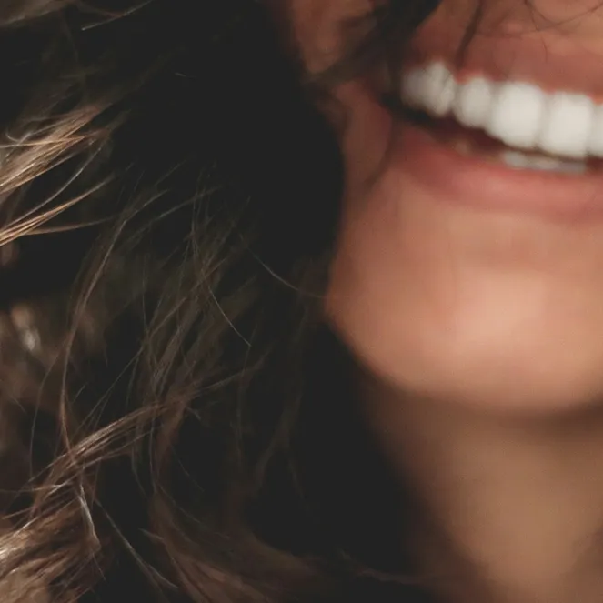 Close-up of a person smiling with white teeth, slightly curled dark hair, and blurred background.