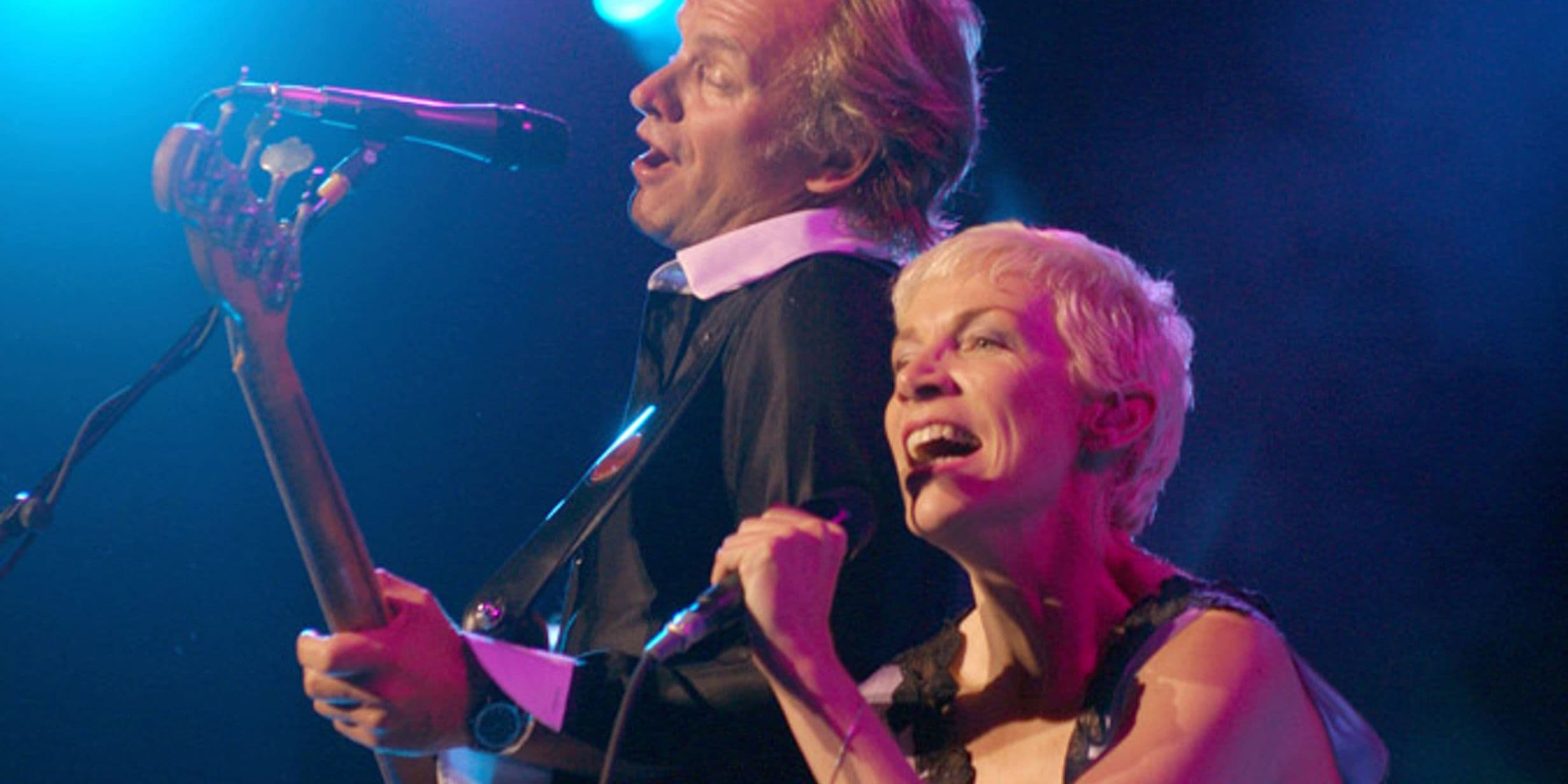 Two performers singing passionately on stage, one playing guitar. Blue stage lights illuminate the scene.