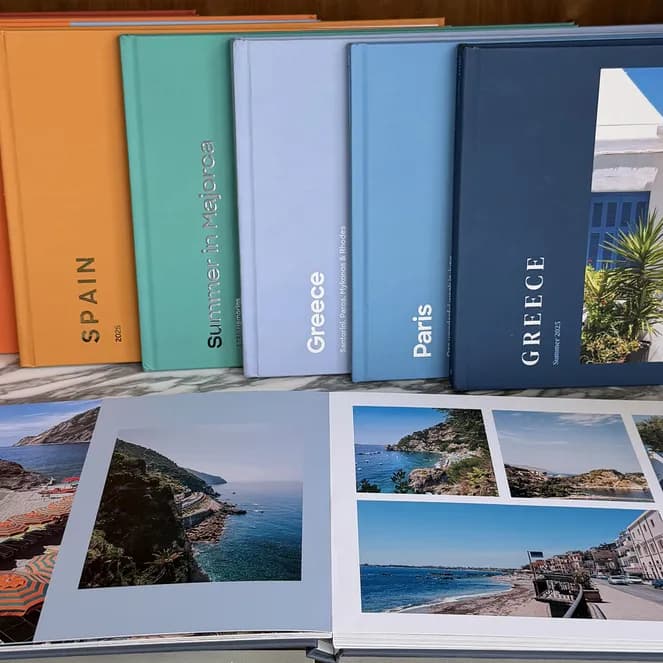 Colorful travel photo albums, including Morocco, Spain, and Greece, on a shelf. One open album displays vibrant beach scenes and coastal views.