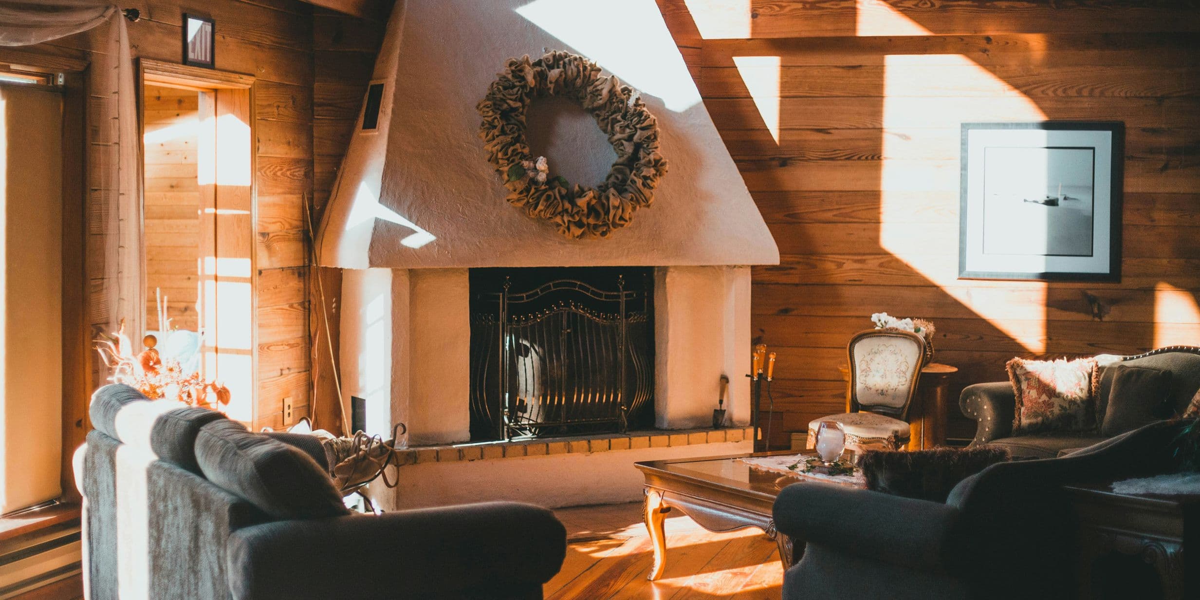 Cozy living room with a white fireplace, wreath, wooden walls, and sunlight streaming through large windows onto sofas and a coffee table.