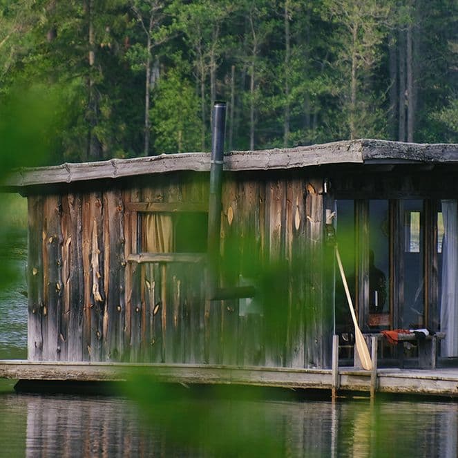Man sitting on the deck of a rustic floating wooden cabin on a calm forest lake, blurred leaves in the foreground.