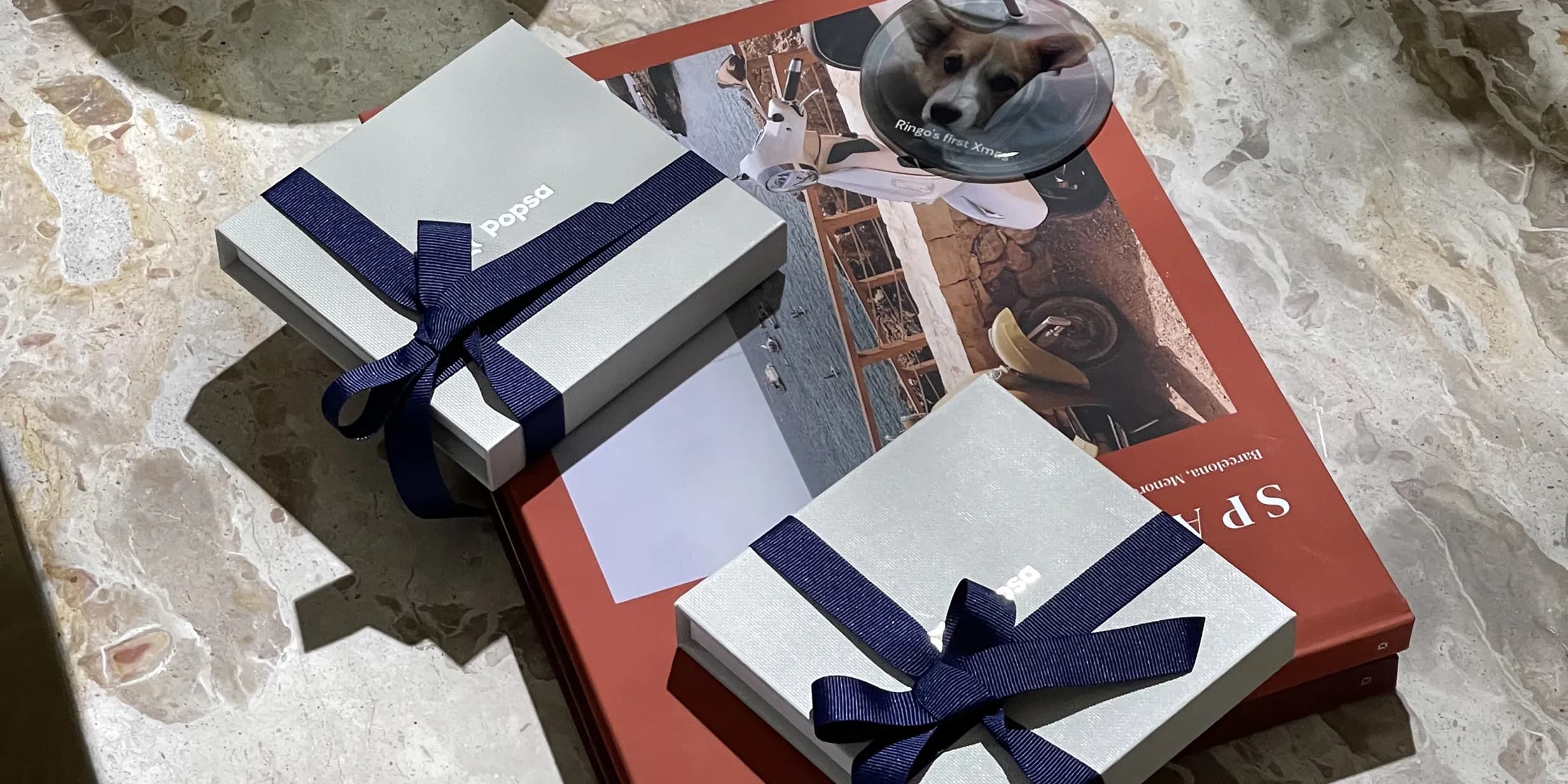 Two gift boxes with navy ribbons on a red book and a coaster with a dog photo on a marble surface.