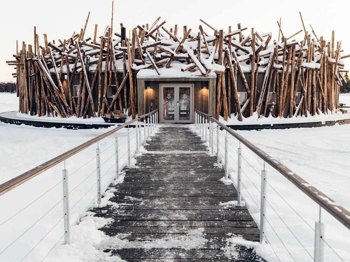 A unique building resembling a large nest of snow-covered logs stands on a vast frozen lake, with a wooden walkway leading to its entrance.