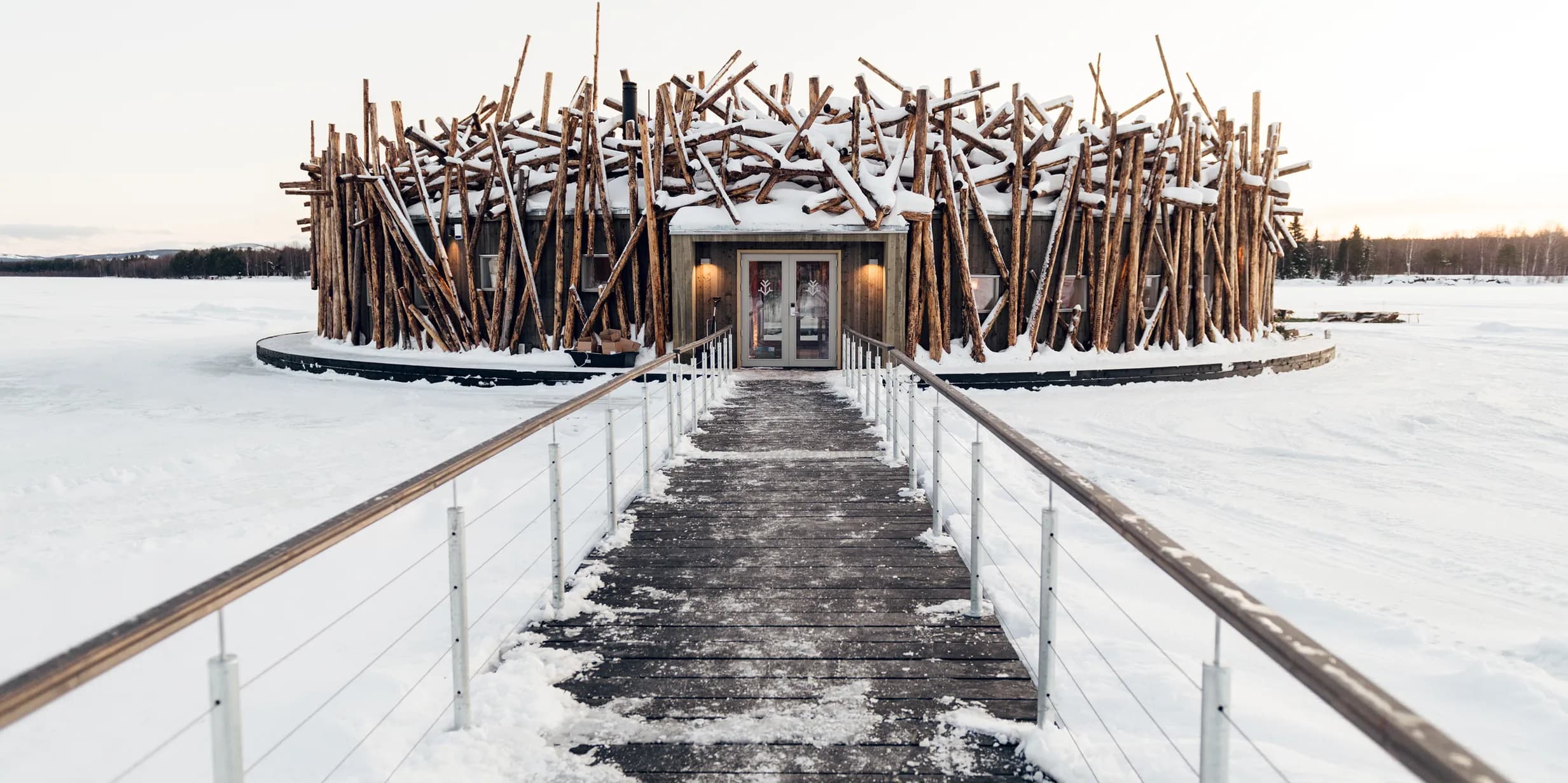 A unique building resembling a large nest of snow-covered logs stands on a vast frozen lake, with a wooden walkway leading to its entrance.