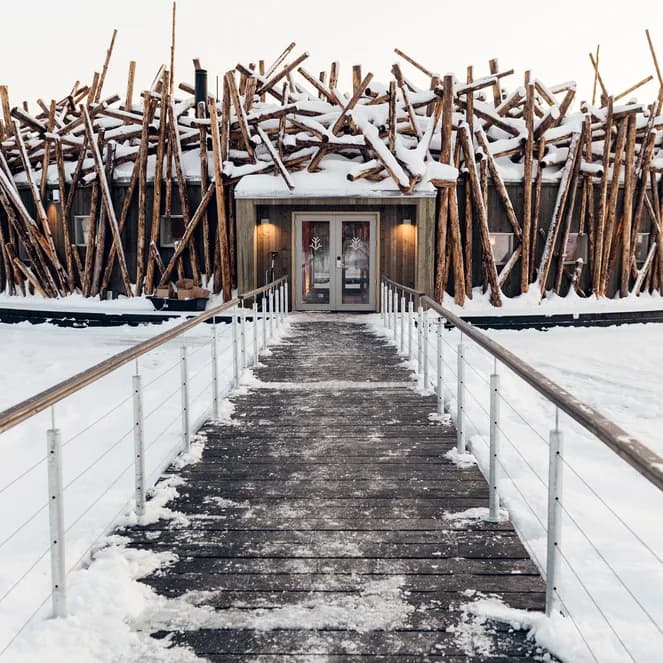 A unique building resembling a large nest of snow-covered logs stands on a vast frozen lake, with a wooden walkway leading to its entrance.