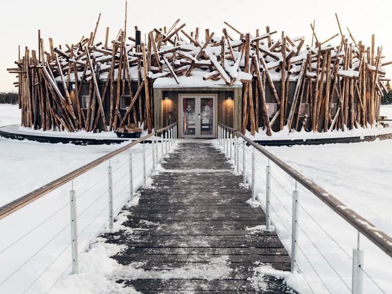 A unique building resembling a large nest of snow-covered logs stands on a vast frozen lake, with a wooden walkway leading to its entrance.