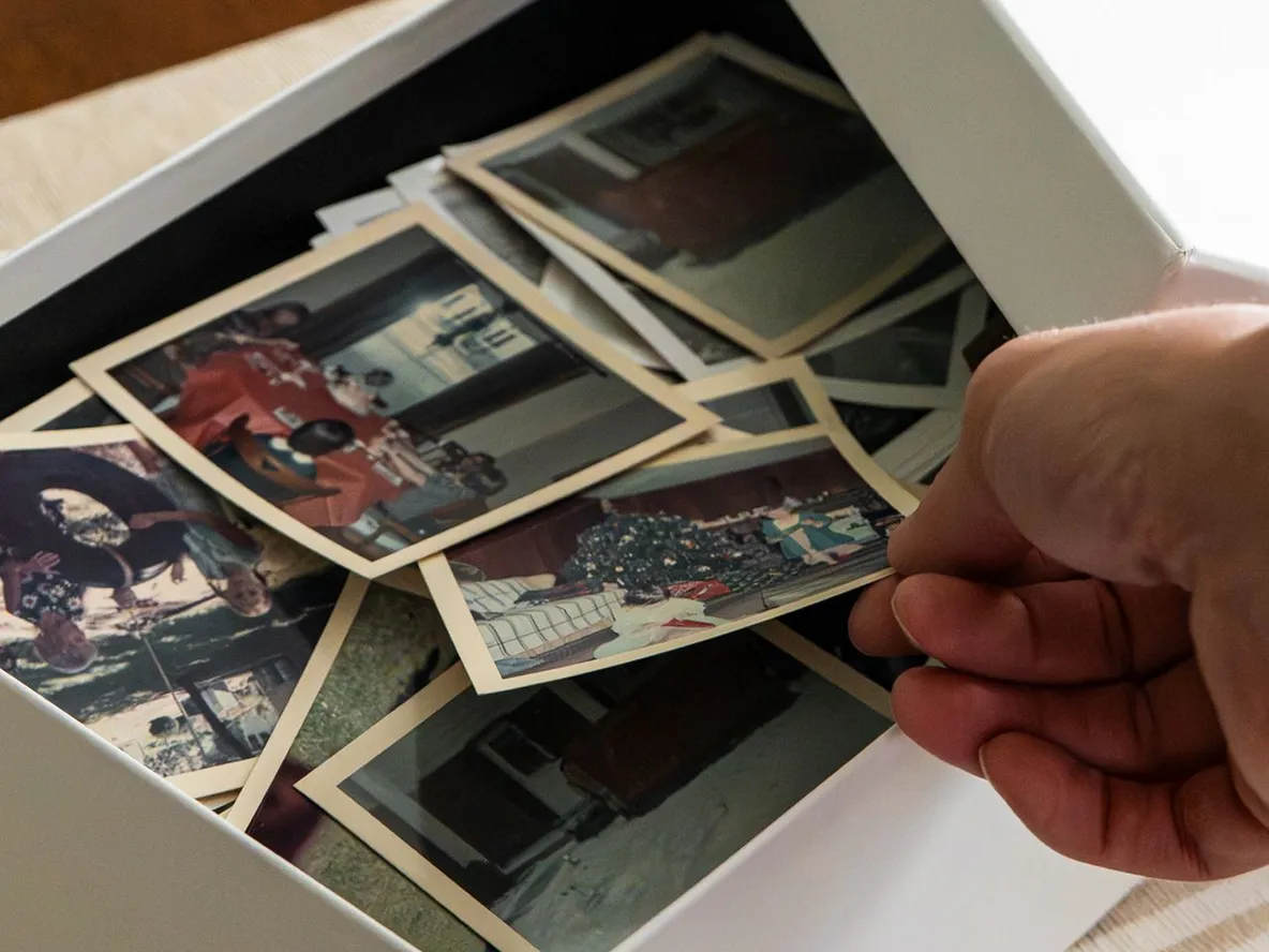 A hand sorting through a box of vintage photographs on a table, featuring various moments and memories.