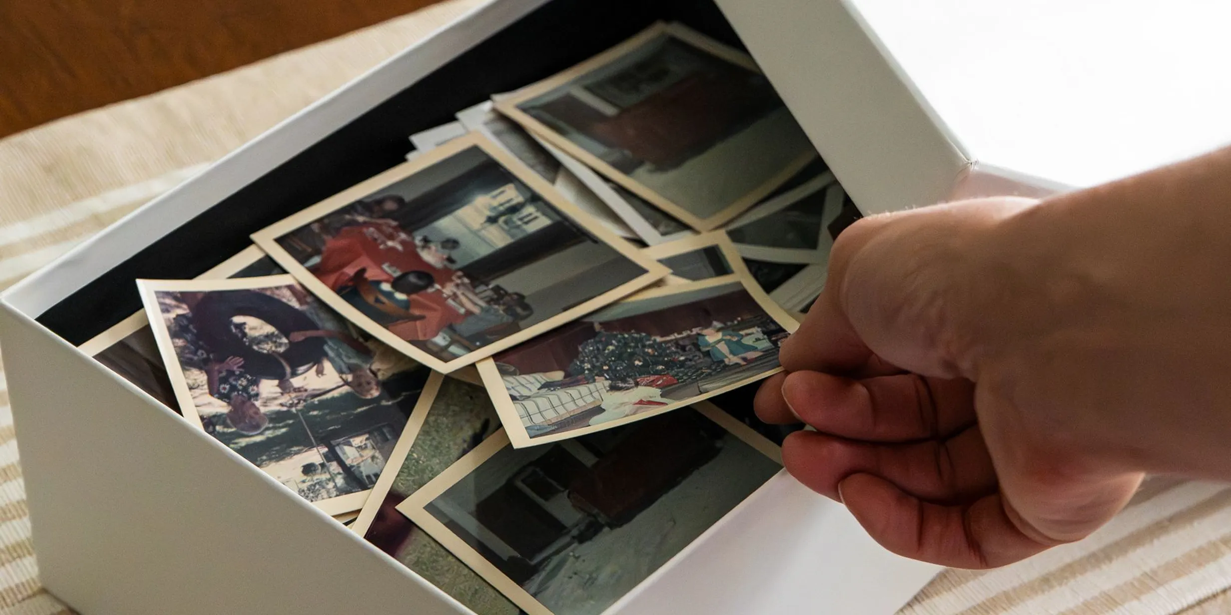 A hand sorting through a box of vintage photographs on a table, featuring various moments and memories.