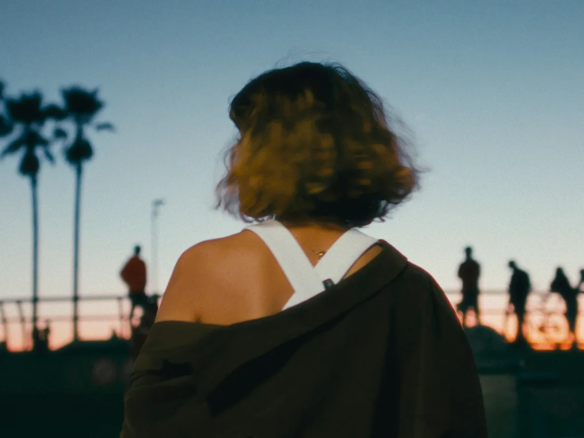 Rear view of person with curly hair, jacket off shoulders, facing a blue and orange sunset with silhouetted palm trees and people.