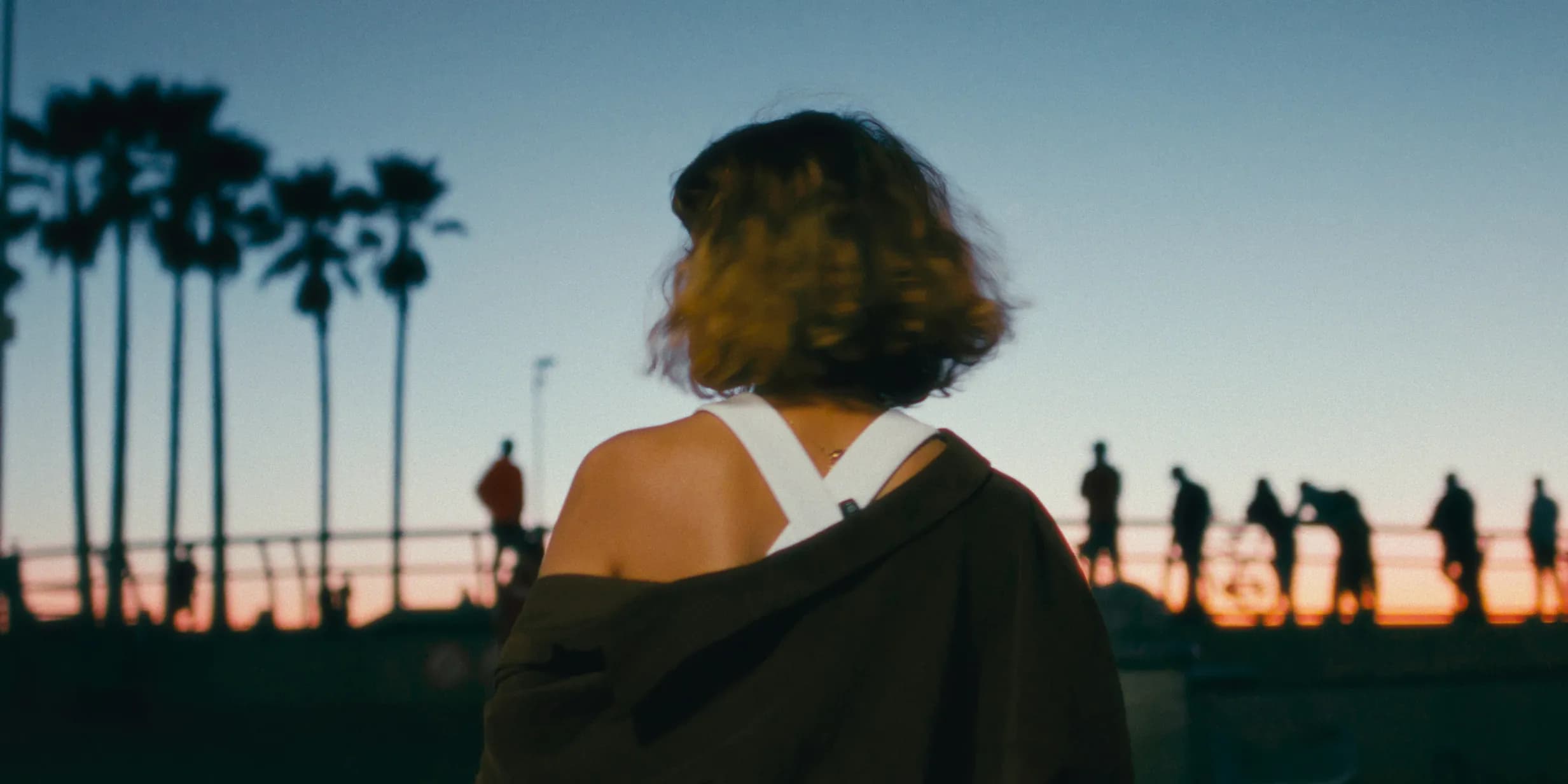 Rear view of person with curly hair, jacket off shoulders, facing a blue and orange sunset with silhouetted palm trees and people.