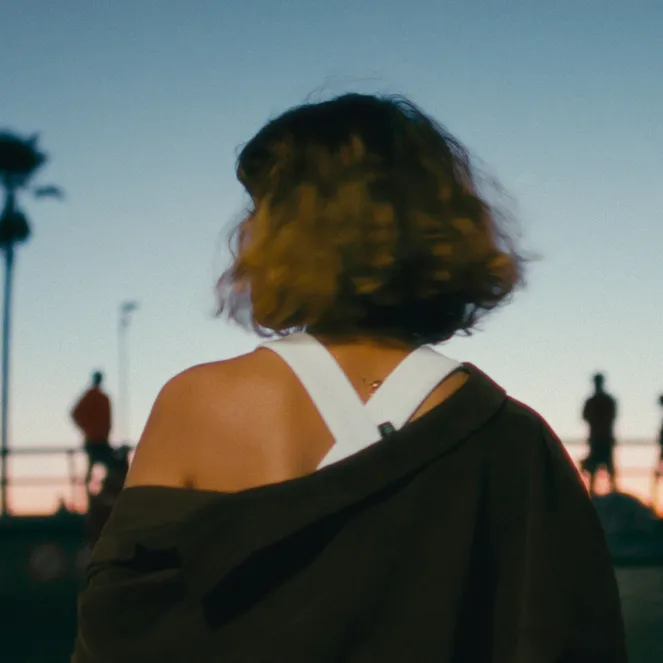 Rear view of person with curly hair, jacket off shoulders, facing a blue and orange sunset with silhouetted palm trees and people.