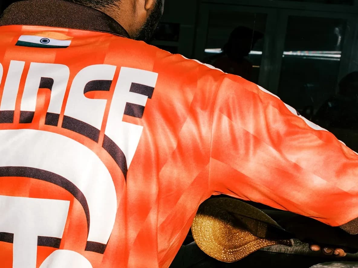 Man wearing an orange shirt with "PRINCE" printed on it, holding a shoe, reflected in a mirror.