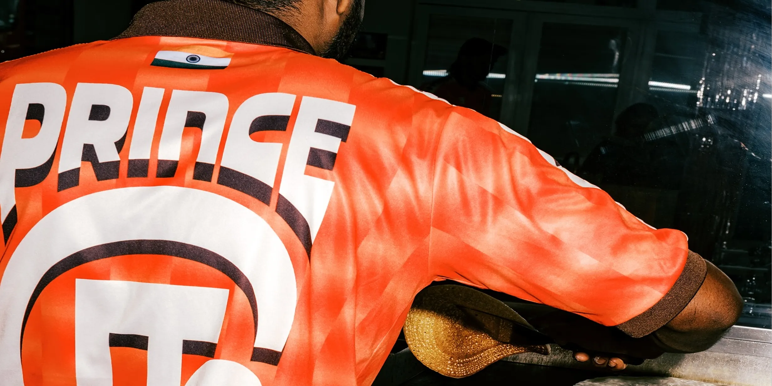 Man wearing an orange shirt with "PRINCE" printed on it, holding a shoe, reflected in a mirror.