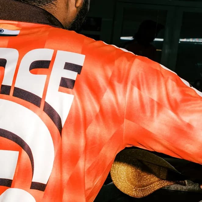 Man wearing an orange shirt with "PRINCE" printed on it, holding a shoe, reflected in a mirror.