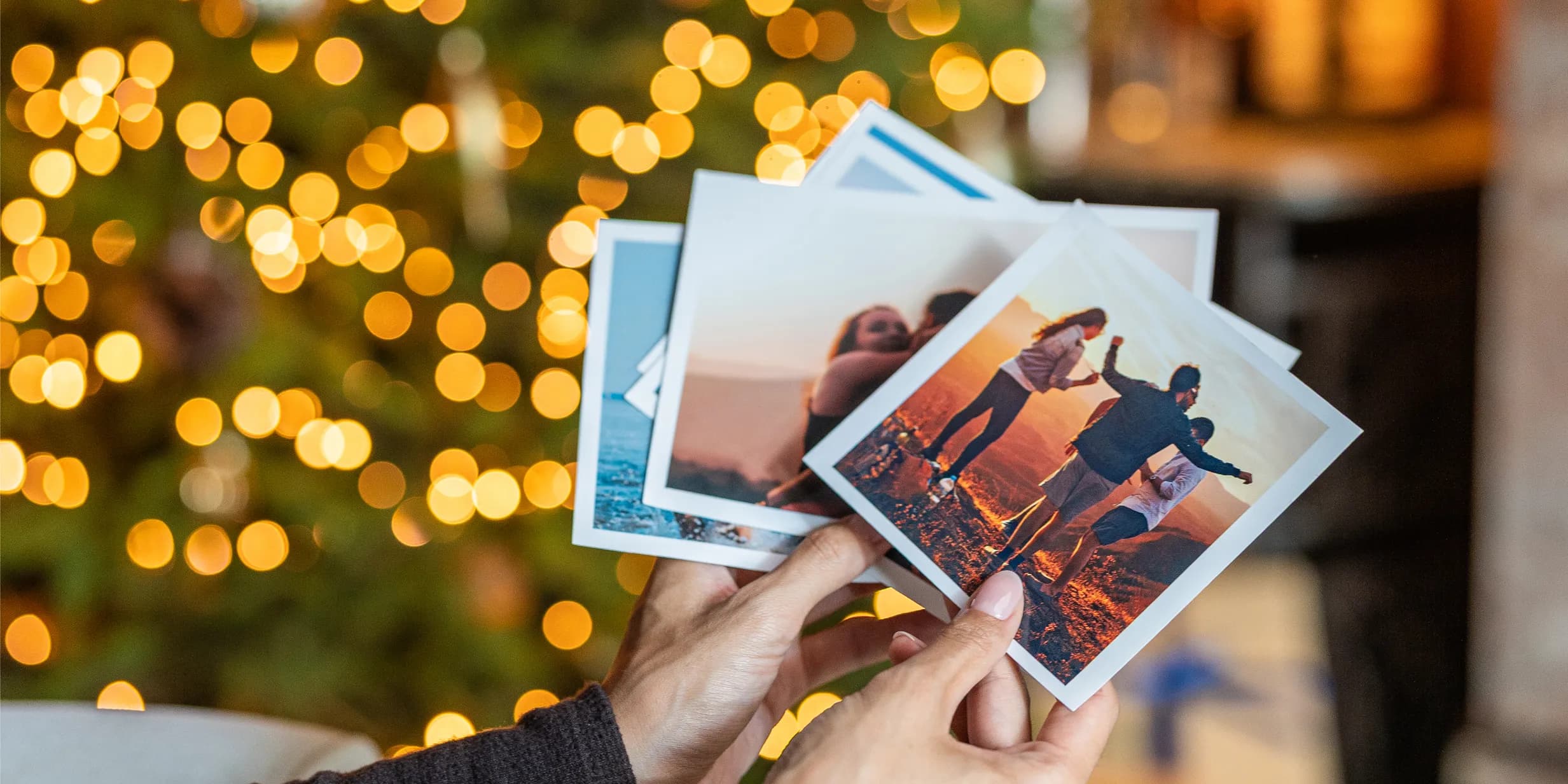 Hands hold several printed photos with white borders against a background of blurred golden Christmas lights.
