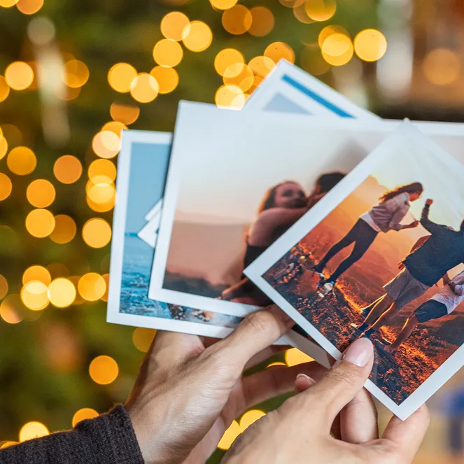 Hands hold several printed photos with white borders against a background of blurred golden Christmas lights.