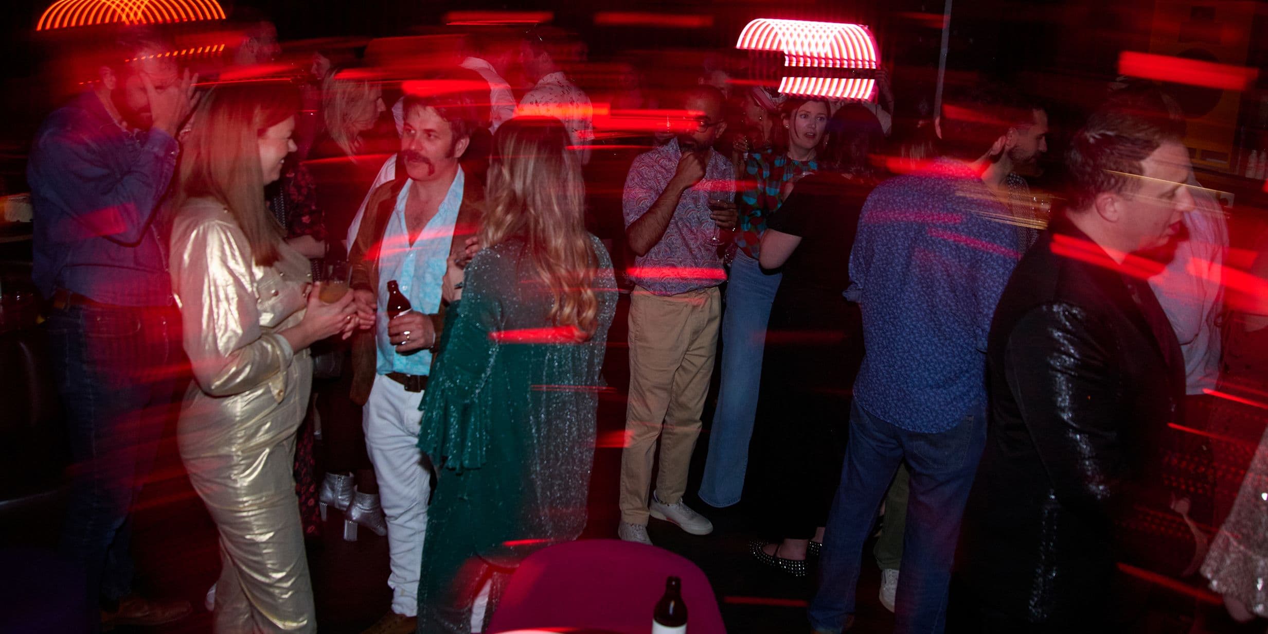 People at a lively party with red light streaks, wearing casual and semi-formal attire, some holding drinks, chatting, and mingling.
