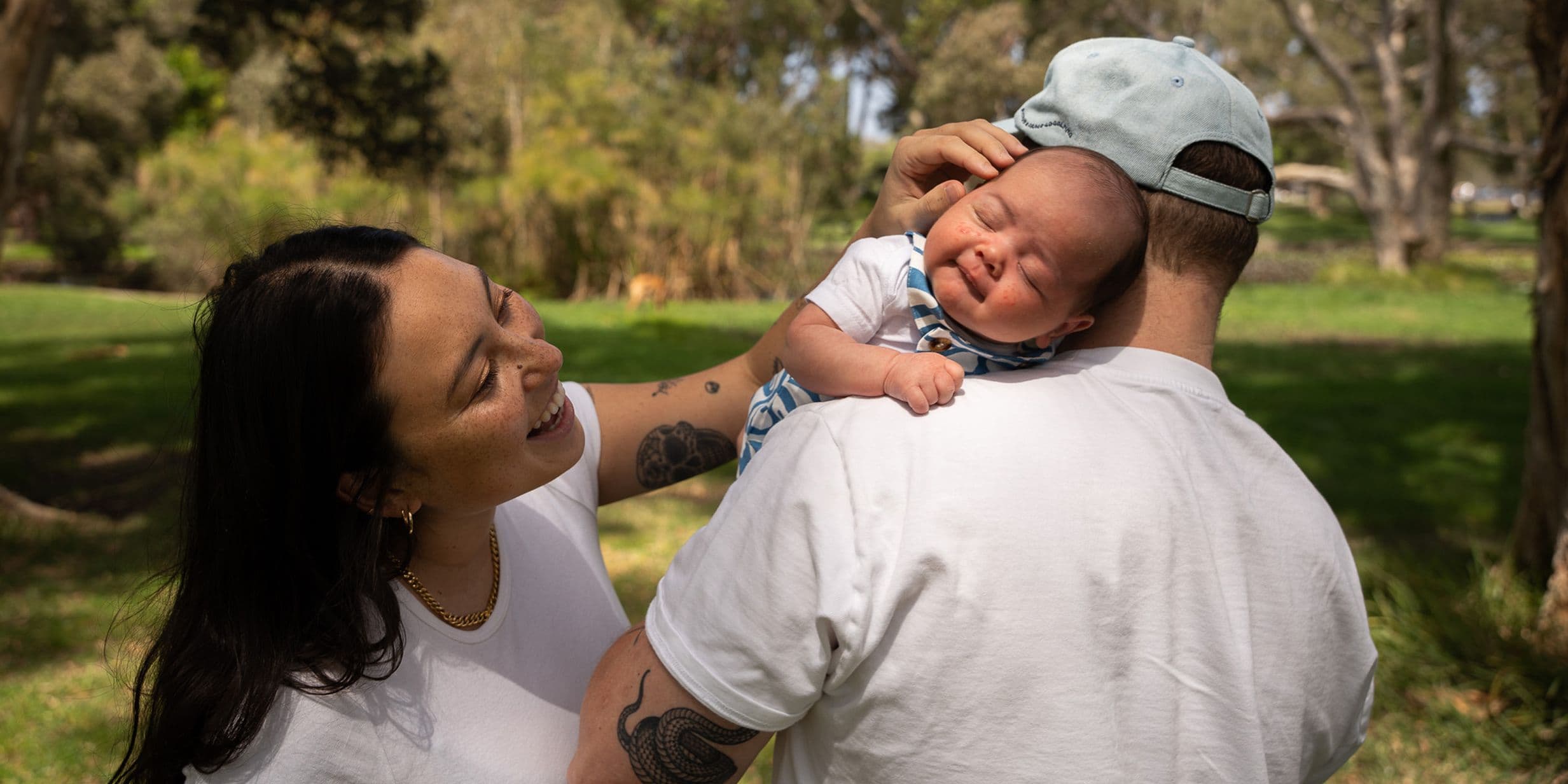 A woman smiles at a baby resting on a man's shoulder outdoors in a park. The baby is wearing a patterned outfit and a cap.