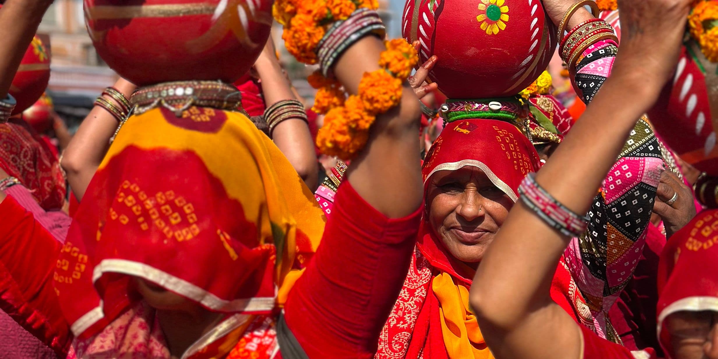 Women in vibrant traditional attire carry decorated pots on their heads during a cultural procession, adorned with floral garlands and bangles.