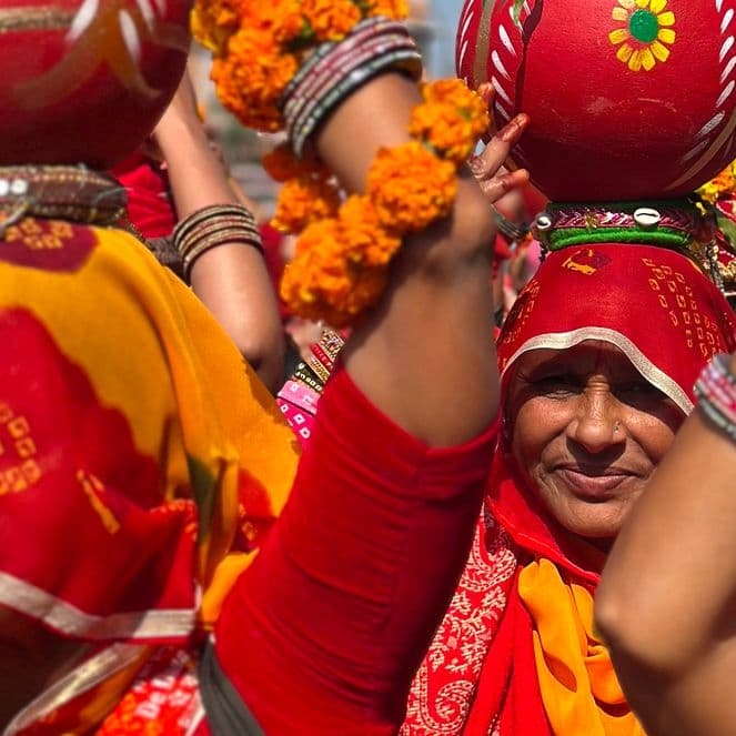 Women in vibrant traditional attire carry decorated pots on their heads during a cultural procession, adorned with floral garlands and bangles.