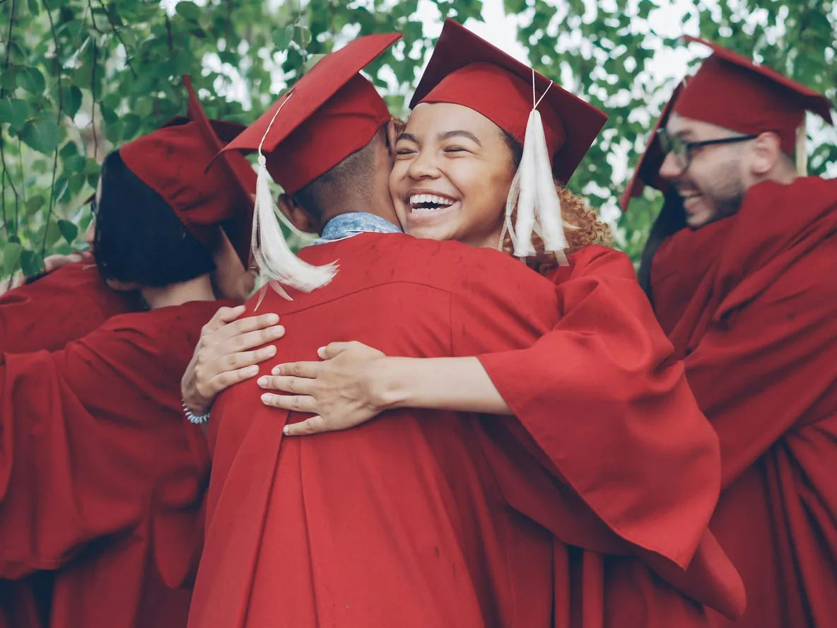 A group of graduates in red caps and gowns joyfully hug each other under leafy trees, celebrating their achievement.