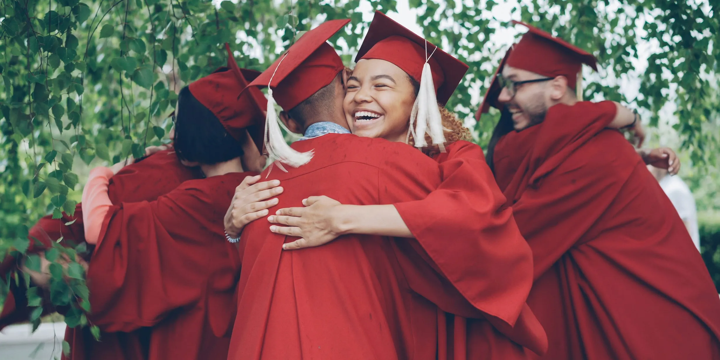 A group of graduates in red caps and gowns joyfully hug each other under leafy trees, celebrating their achievement.