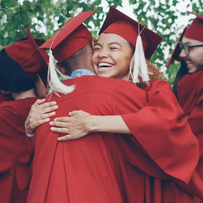A group of graduates in red caps and gowns joyfully hug each other under leafy trees, celebrating their achievement.