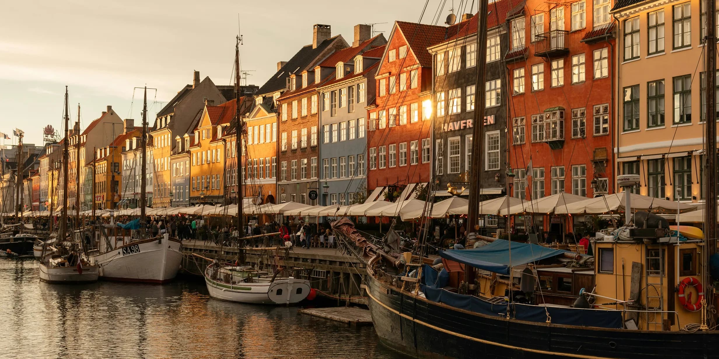 Colorful buildings and moored boats along a canal in Nyhavn, Copenhagen, under warm, golden evening light, with lively outdoor cafes.