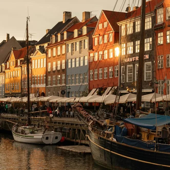 Colorful buildings and moored boats along a canal in Nyhavn, Copenhagen, under warm, golden evening light, with lively outdoor cafes.