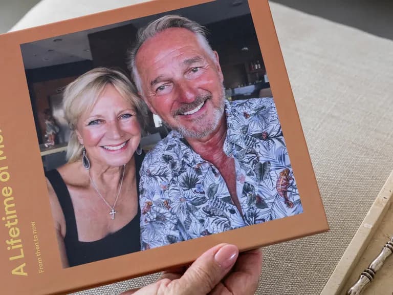 A person holds a photo album titled "A Lifetime of Memories" with a smiling couple on the cover. Nearby is a lit candle on a tray.