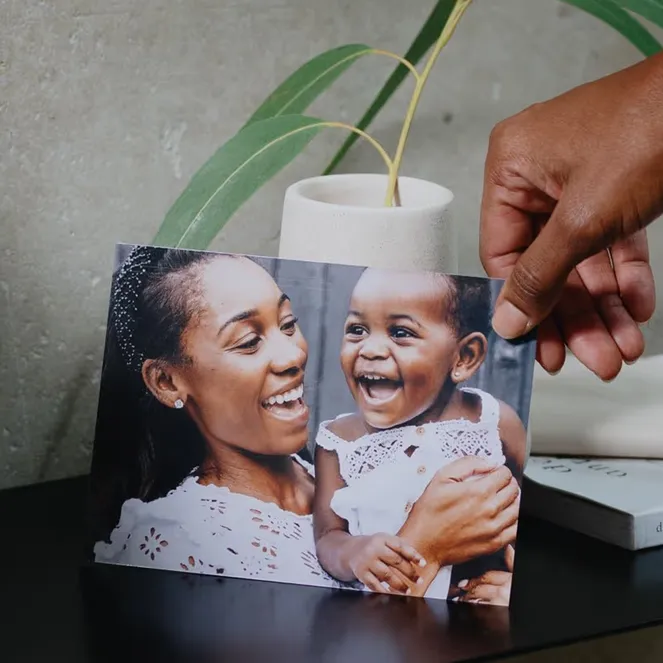 Dark-skinned hand places a photo of a joyful Black mother smiling with her laughing baby onto a dark shelf.
