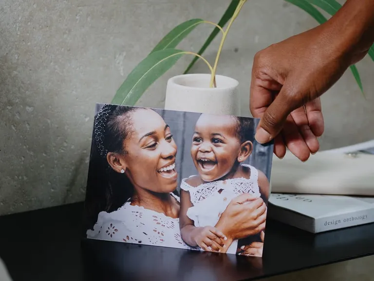 Dark-skinned hand places a photo of a joyful Black mother smiling with her laughing baby onto a dark shelf.