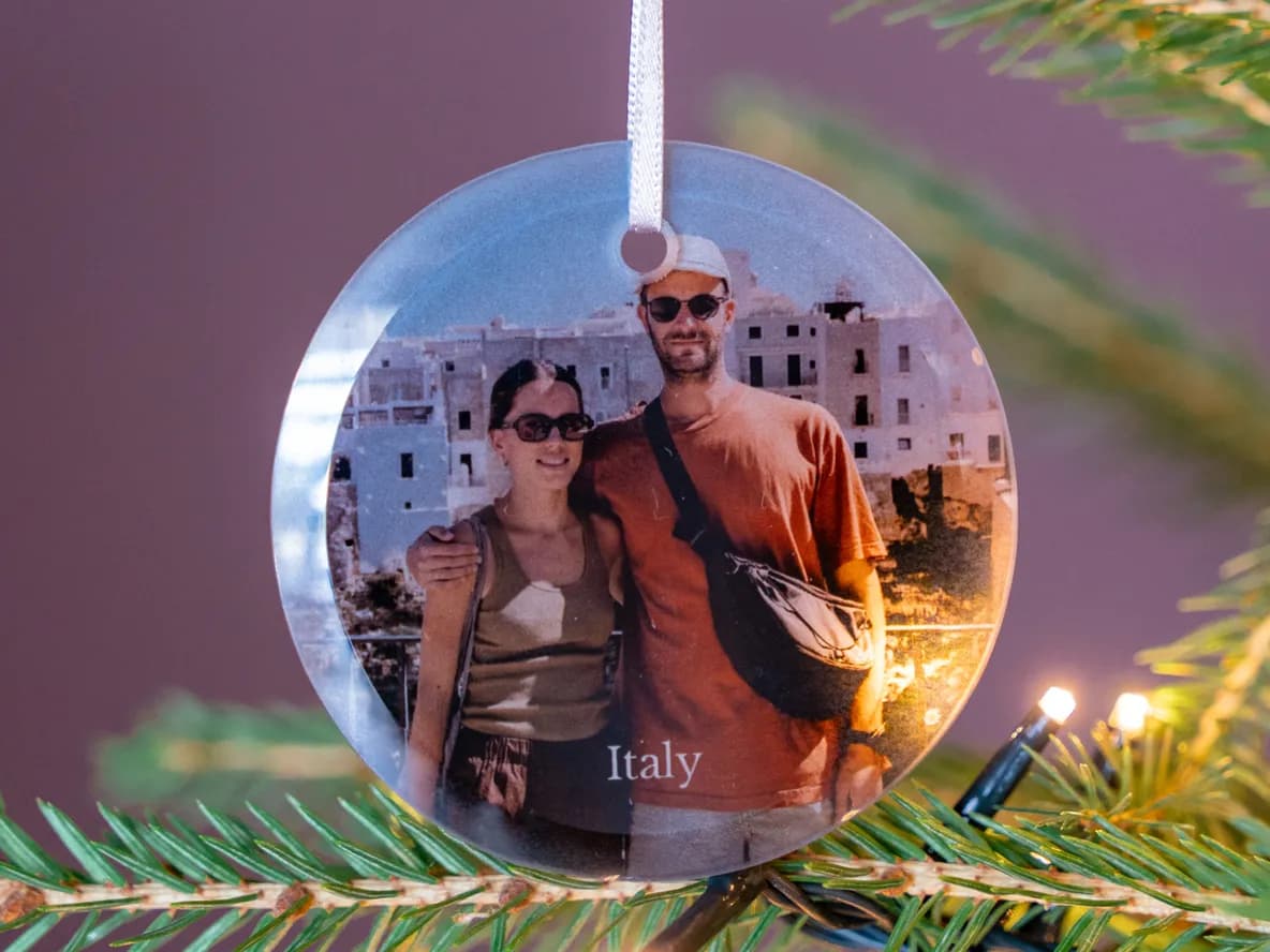 A circular Christmas ornament featuring a photo of a smiling couple in Italy hangs on a green Christmas tree with twinkling lights.