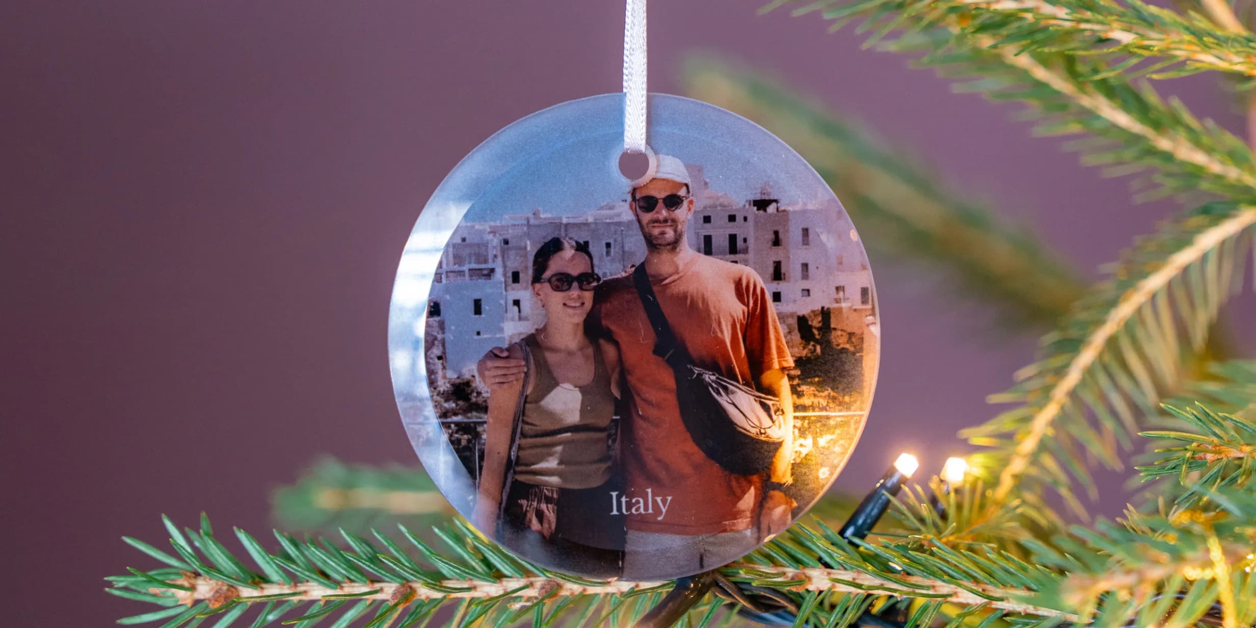 A circular Christmas ornament featuring a photo of a smiling couple in Italy hangs on a green Christmas tree with twinkling lights.