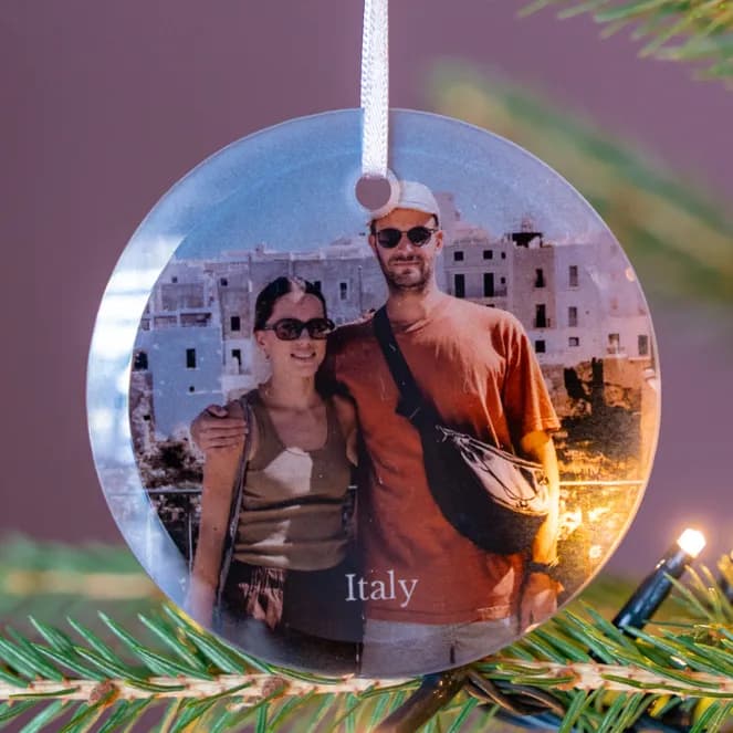 A circular Christmas ornament featuring a photo of a smiling couple in Italy hangs on a green Christmas tree with twinkling lights.