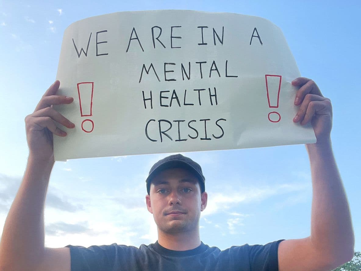 Person holding a sign reading "We are in a mental health crisis" with exclamation marks, against a blue sky background.