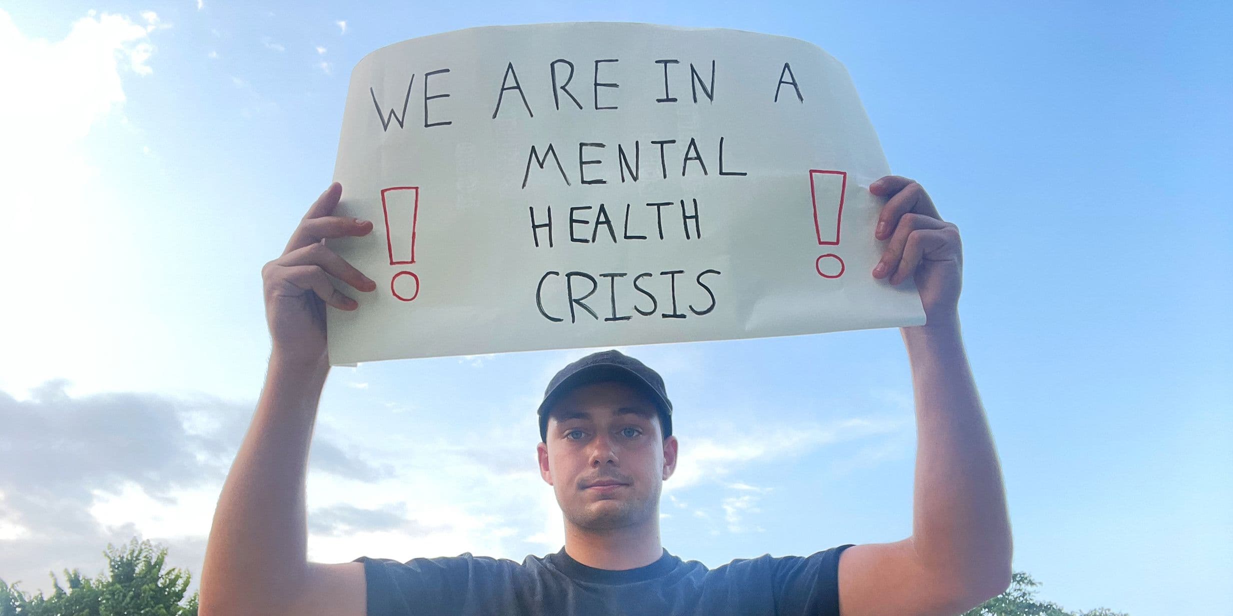 Person holding a sign reading "We are in a mental health crisis" with exclamation marks, against a blue sky background.
