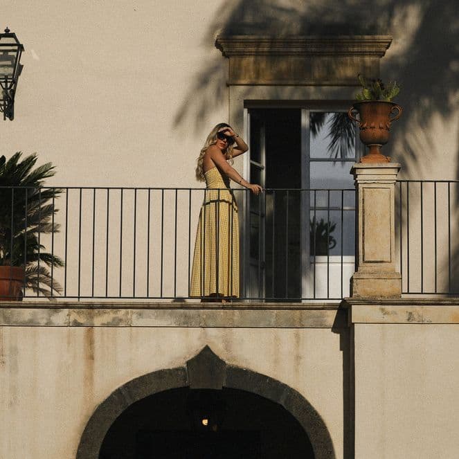 A person in a yellow dress stands on a balcony with potted plants, under the shadow of palm trees, against a beige building.