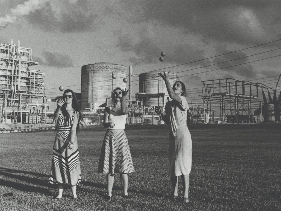 Three women in dresses stand on grass before a large industrial power plant with smokestacks and electrical towers.