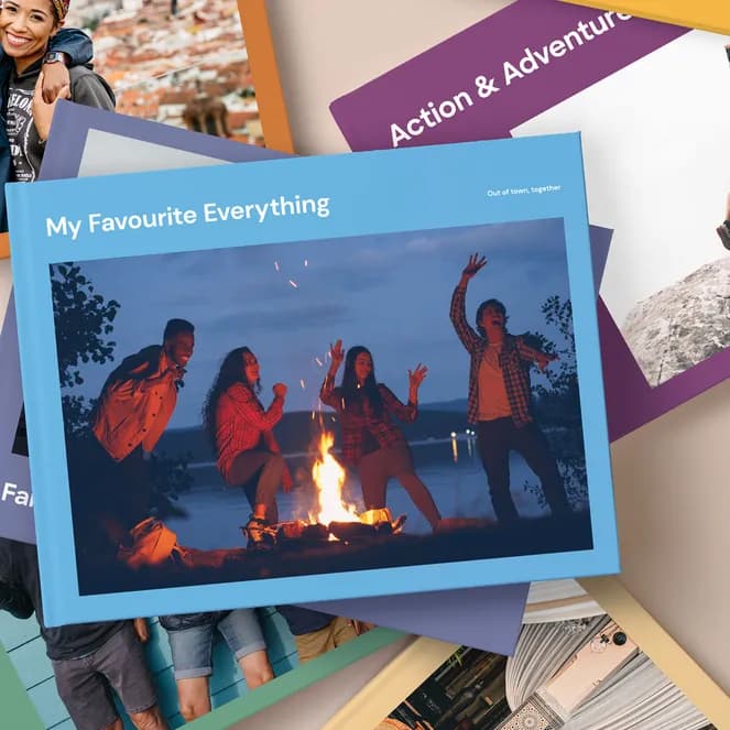 Stack of photo books with various covers, including a couple, a campfire scene, and people jumping on rocks, against a beige background.
