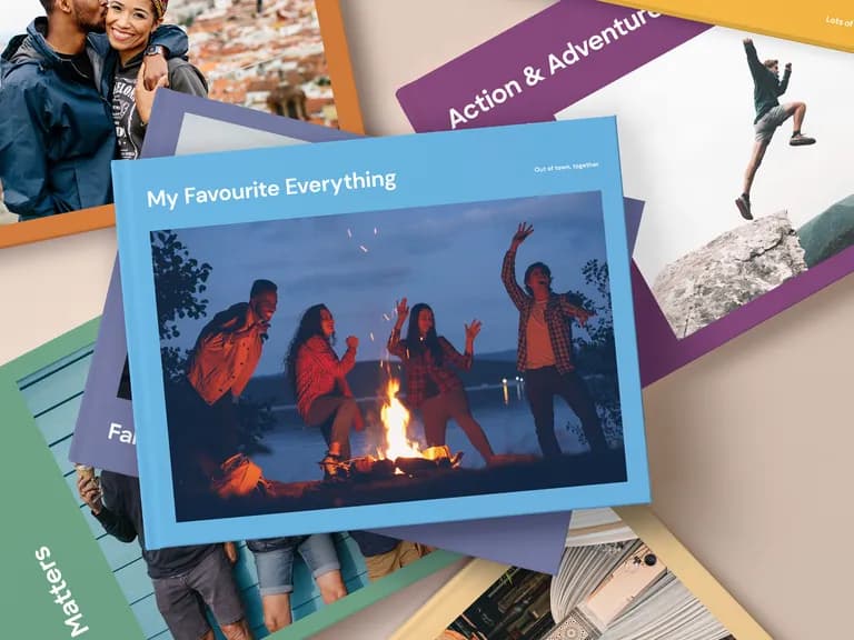 Stack of photo books with various covers, including a couple, a campfire scene, and people jumping on rocks, against a beige background.