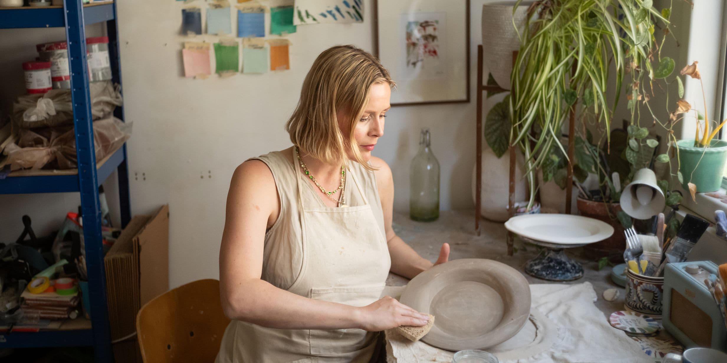 A woman in an apron shapes a clay bowl in a pottery studio with plants and art supplies surrounding her.